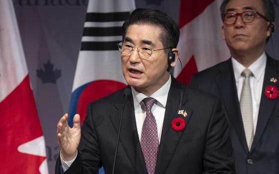 A South Korean man wearing a suit and a red flower pin speaks into a microphone with South Korean and Canadian flags behind him.