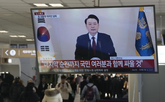 People walk beneath a mounted television screen showing the South Korean president wearing a suit and speaking in front of the South Korean flag.