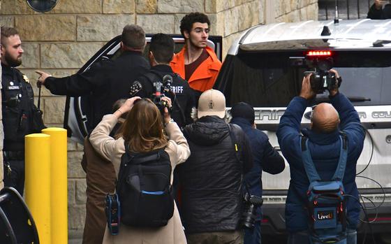 The suspect in the murder of a health insurance company CEO is emerges from a police van while surrounded by police and reporters.