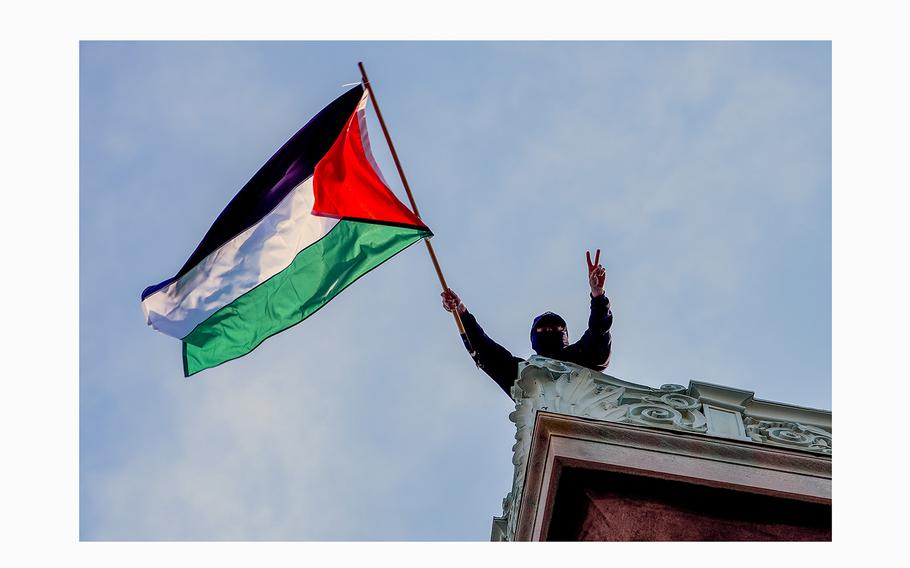 The head and shoulders of a masked student protester in black on top of a building are seen from below, with the student holding a Palestinian flag and making a peace sign.