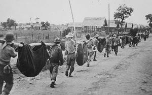 A black-and-white photo of a line of men carrying poles that hold wrapped corpses. 