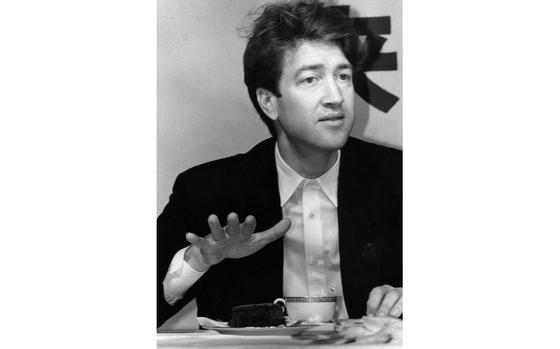 A young David Lynch raises a hand to chest level while seated at a table in front of a plate of food and a cup.