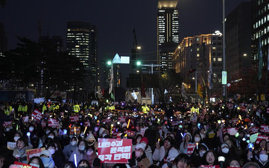 People hold signs outdoors at nighttime in a city with skyscrapers.