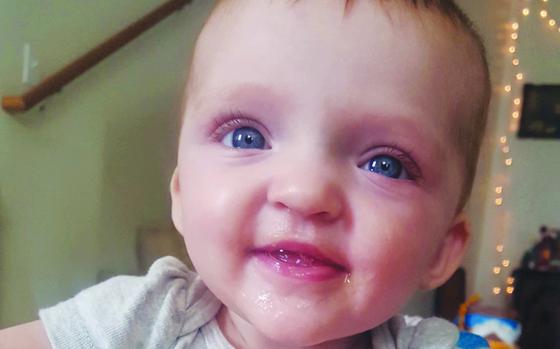 A baby girl with big blue eyes smiles with twinkle lights behind her.