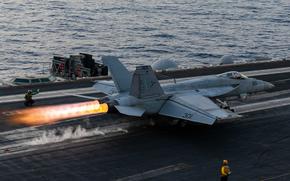 A line of fire shoots out from the back of a military fighter jet as it prepares to take off from the deck of an aircraft carrier.