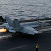 A line of fire shoots out from the back of a military fighter jet as it prepares to take off from the deck of an aircraft carrier.