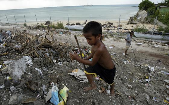 A shirtless and barefoot boy scavenges among trash near the beach/sea.