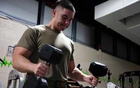 A military member in camouflage pants and olive green shirt lifts dumbbells with each hand while exercising in a gym.