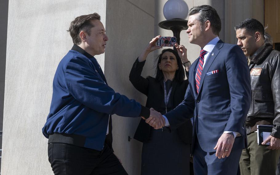 Pete Hegseth and Elon Musk shake hands on stone steps outside the Pentagon as people behind them use their phones to take photos.