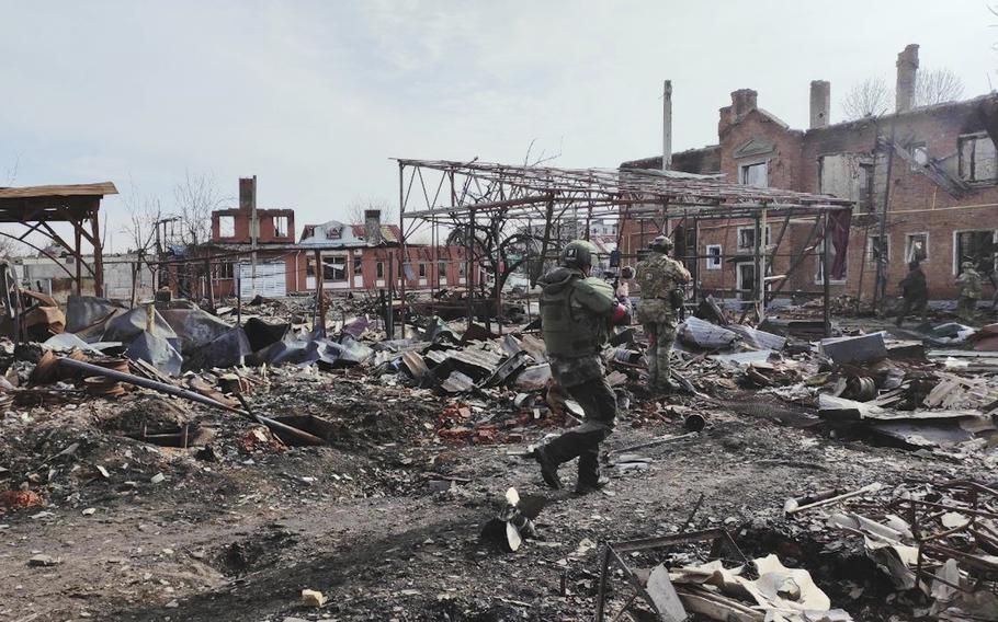 Two Russian soldiers  in the midground walk past the remnants of destroyed buildings while other soldiers are seen in the background.