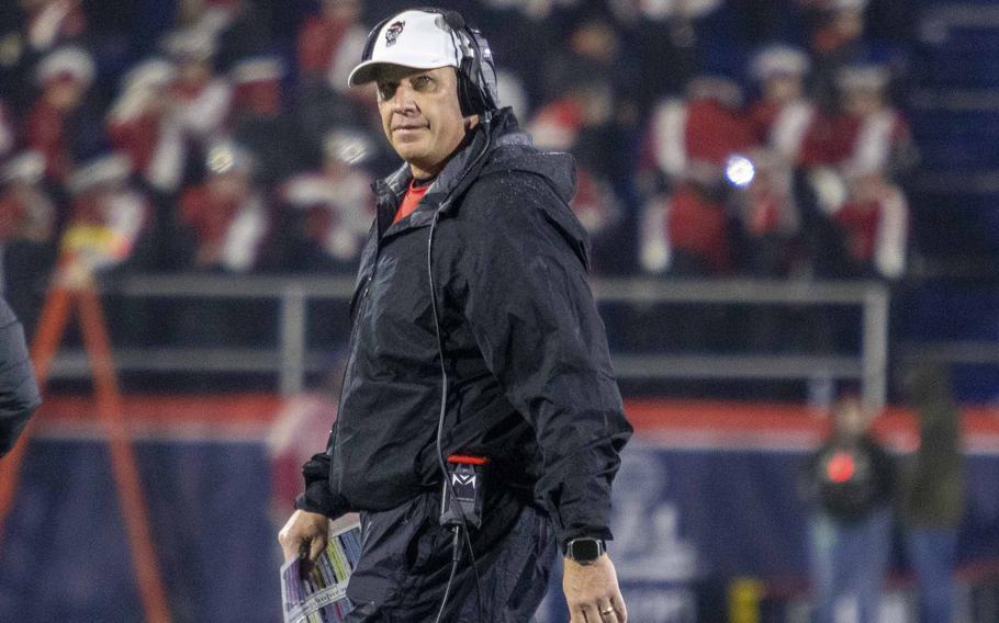 A man wearing a track suit and a baseball hat and headset walks along the side of a football field.