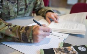 A person in uniform holds open a book while writing in a notebook. 