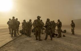 A group of soldier in combat gear stand next to a desert road under a low sun.