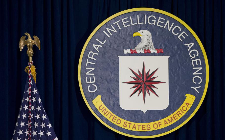 The yellow bordered blue seal of the central intelligence agency is shown against a black background beside an American flag.