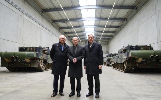 Three men in suits and overcoats stand in a warehousee with rows of tanks behind them.