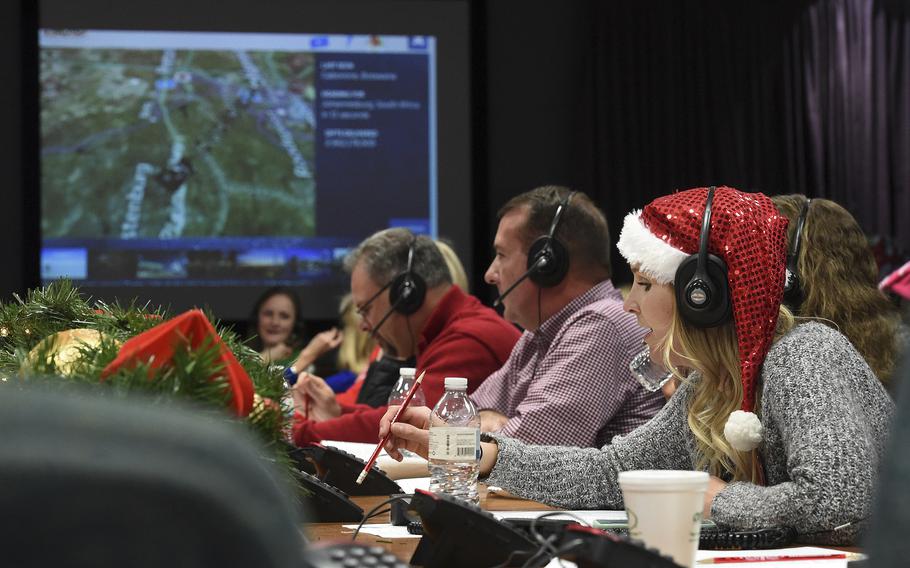 Volunteers in Santa hats and radio headsets sit at a long table in front of a digital display screen.