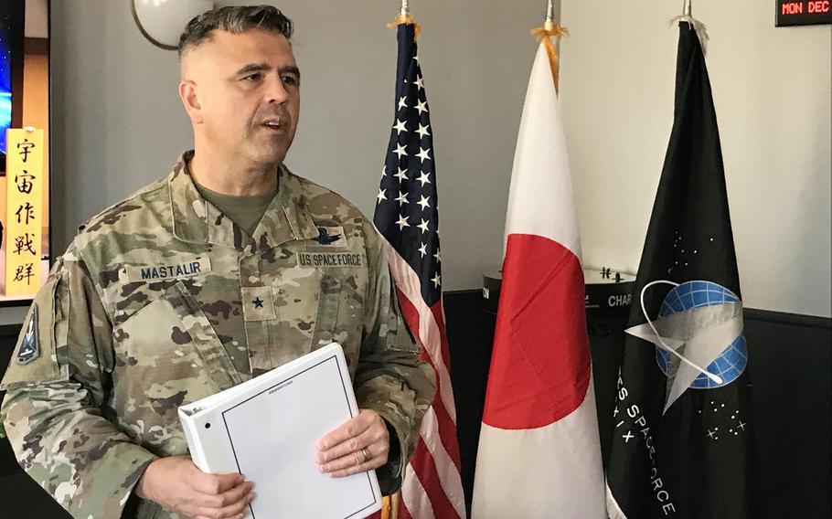 A brigadier general speaks with reporters in front of a U.S. flag, a Japanese flag and a Space Force flag.