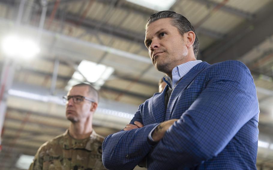 Hegseth standing with his arms crossed and a soldier to his right.