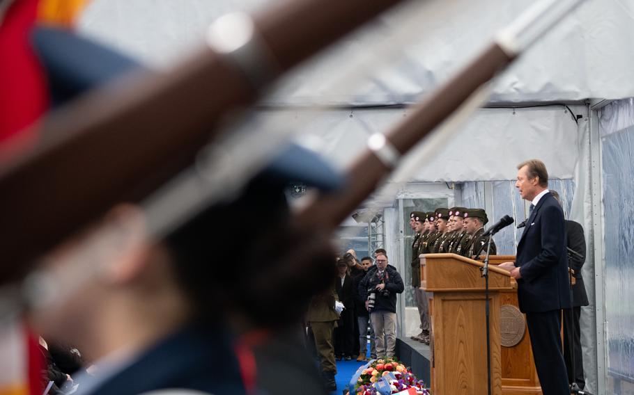 Out-of-focus rifles closer to the camera cover the left half of the image while Luxembourg’s Grand Duke Henri is in-focus on the image’s right, farther away from the camera.