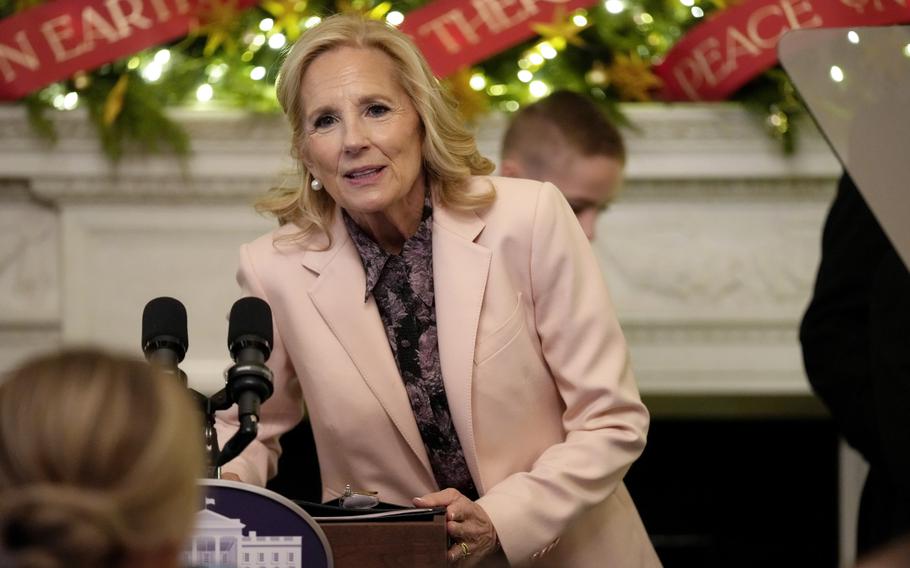 Jill Biden speaks indoors at a podium with holiday decorations behind her.