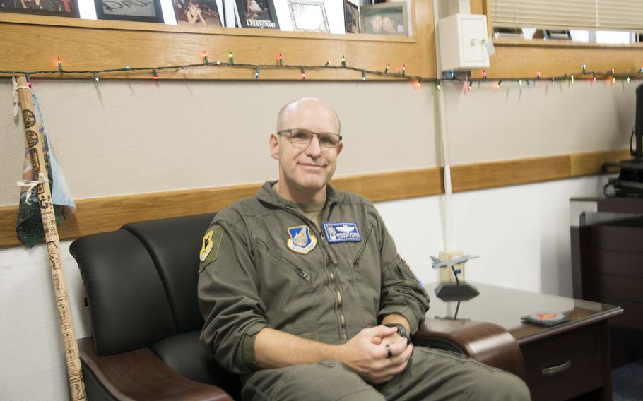 A man wearing a flight suit sits on a leather chair with a stamped hiking stick next to him resting next to him and twinkle lights strung above him.