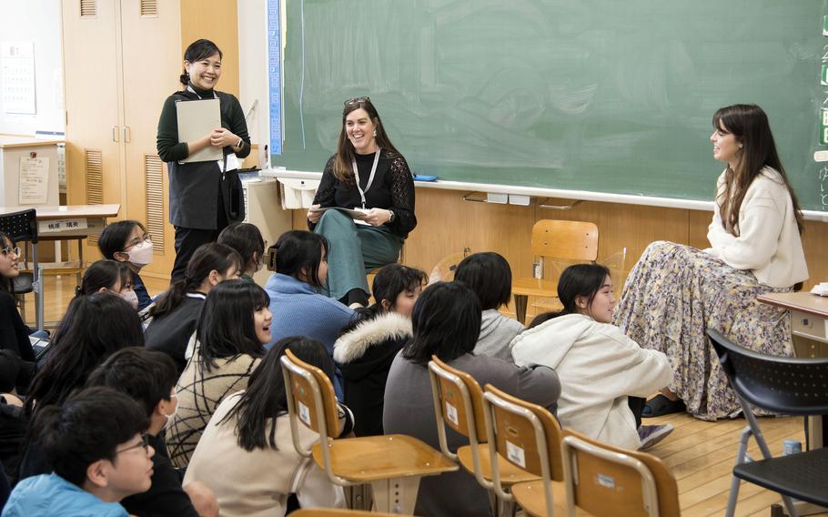 Two American teachers sit in chairs at the front of a classroom and speak to a group of Japanese students seated on the floor.