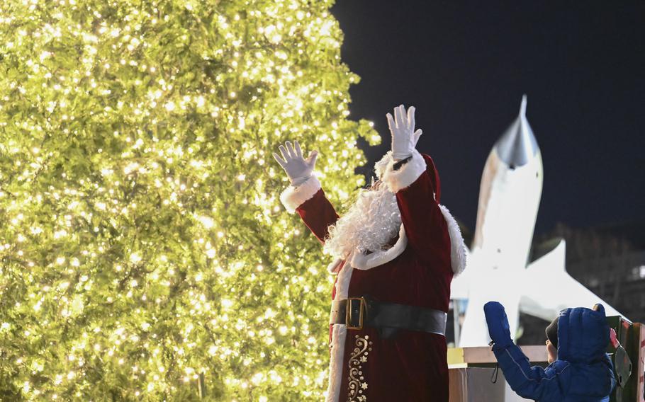 An Air Force officer dressed as Sant Claus raises his arms in celebration in front of the the just-lit Christmas tree.