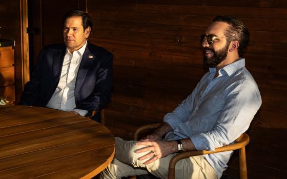 Two men in button-down shirts speak while sitting at a wooden table.