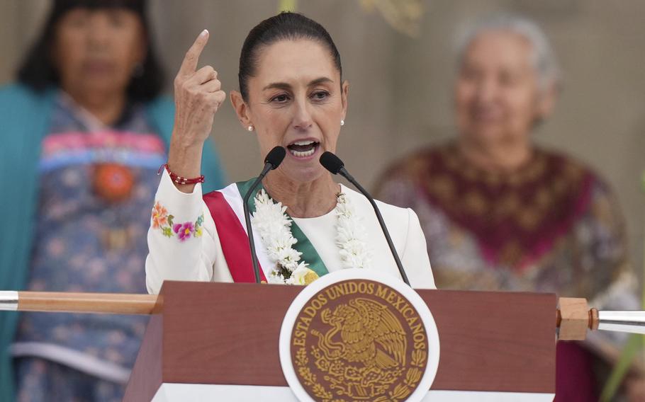 Mexican President Claudia Sheinbaum gestures with her hand as she stands at a podium and speaks into microphones.