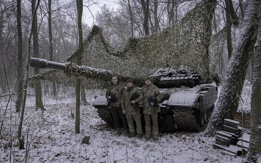 Three Ukraine solider stand in front of a tank, with a camouflage net above and snow on the ground.