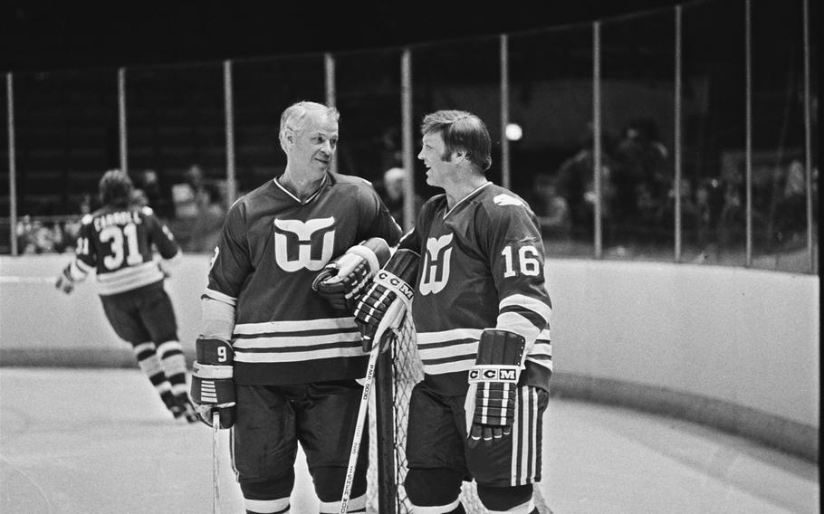 Bobby Hull and another hockey player, both wearing uniforms, lean on their hockey sticks and chat on the ice.