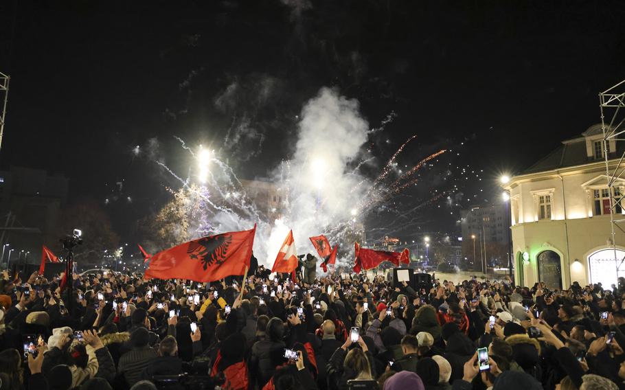 Fireworks explode in the center of the photo while a mass of revelers with flags and phones out take up most of the bottom third.