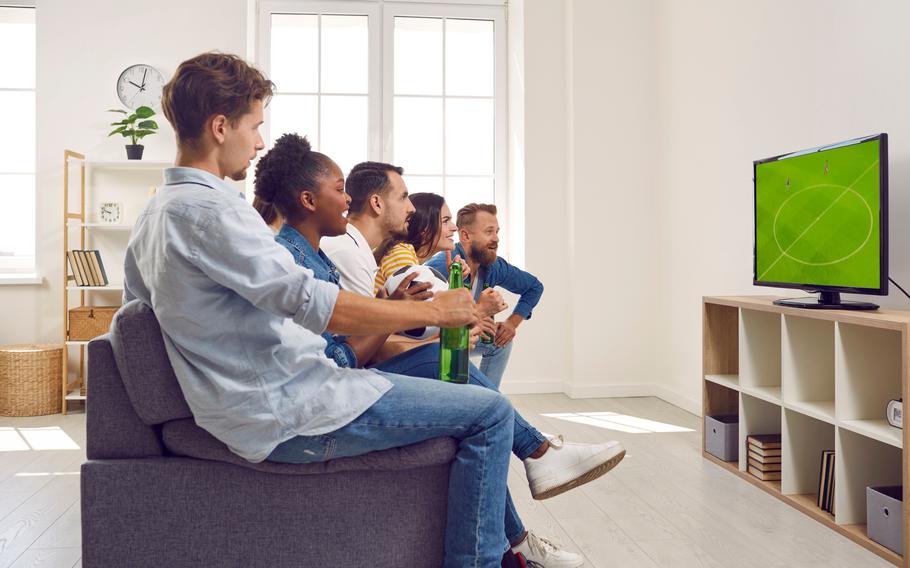 Young guys and girls watching soccer football game on TV together sitting on couch in living room