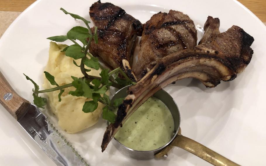 Three lamb chops sit on a white plate with a green sauce, mashed potatoes and green sprigs.