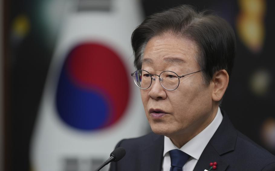 A man in a suit with glasses and a small South Korea flag lapel pin speaks in front of a South Korean flag.