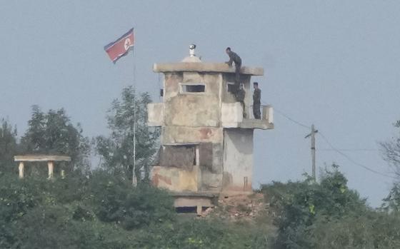 Soldiers stand on a balconey of a military guard post while a North Korean flag flutters in the wind next to it.