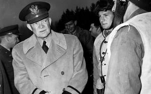 A black-and-white, 1950s-era image of Dwight Eisenhower in a military overcoat speaking with troops in combat gear.