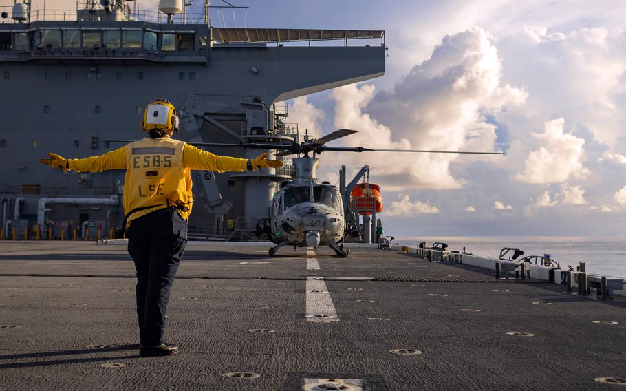 An air traffic controller stands in front of a helicopter as it prepares to take off from the deck of a ship.