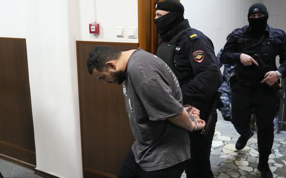 A man with his head bowed and hands cuffed behind him is led into a courtroom by a police officer.