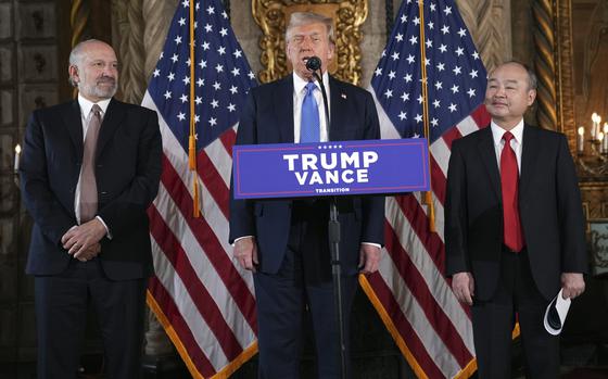 Donald Trump stands at a podium in front of U.S. flags and speaks into a microphone while flanked by two men in dark suits.