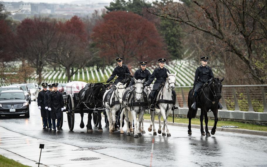 Soldiers riding horses pull a casket on a wagon as other troops walk along side of the wagon and cars follow.