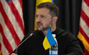 Volodymyr Zelenskyy sits at a microphone with a small Ukrainian flag on the table in front of him.