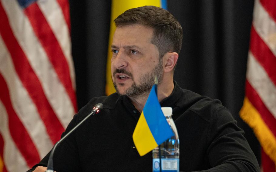 Volodymyr Zelenskyy sits at a microphone with a small Ukrainian flag on the table in front of him.