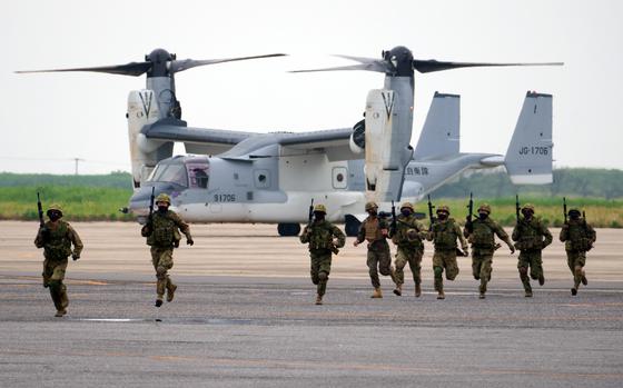 Soldiers in combat gear and holding machine guns run across a tarmac in front of an Osprey aircraft.