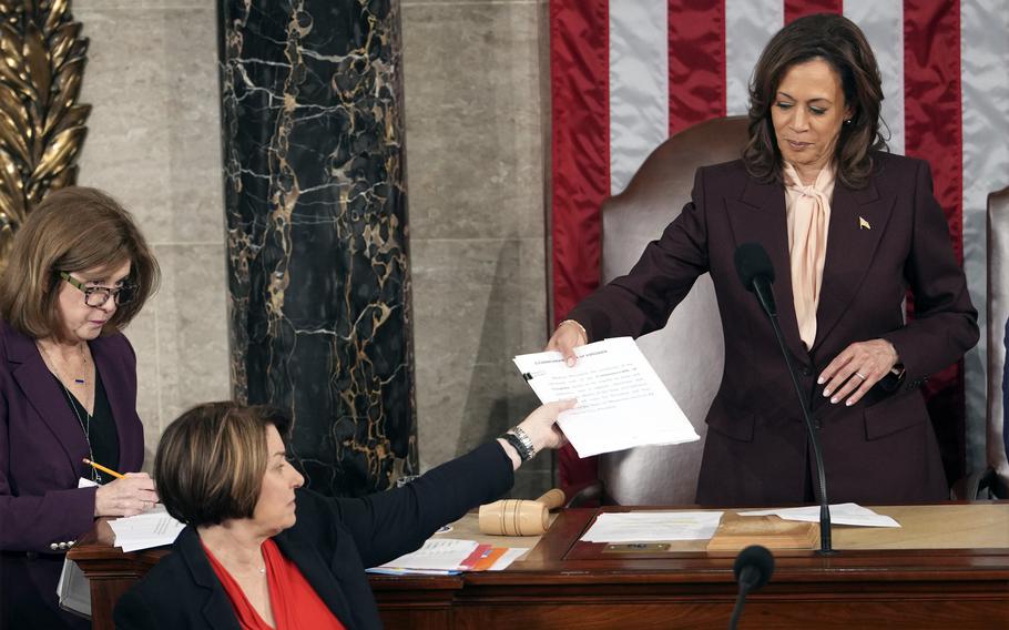 Kamala Harris, with the flag in the background, hands a document to Amy Klobuchar in the House chamber.