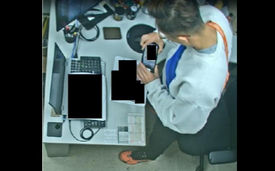 Surveillance video footage of a man, seen from above, using his phone to photograph documents on a table.