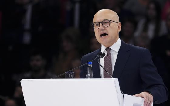 A bald man with a suit and glasses holds a white podium while speaking.