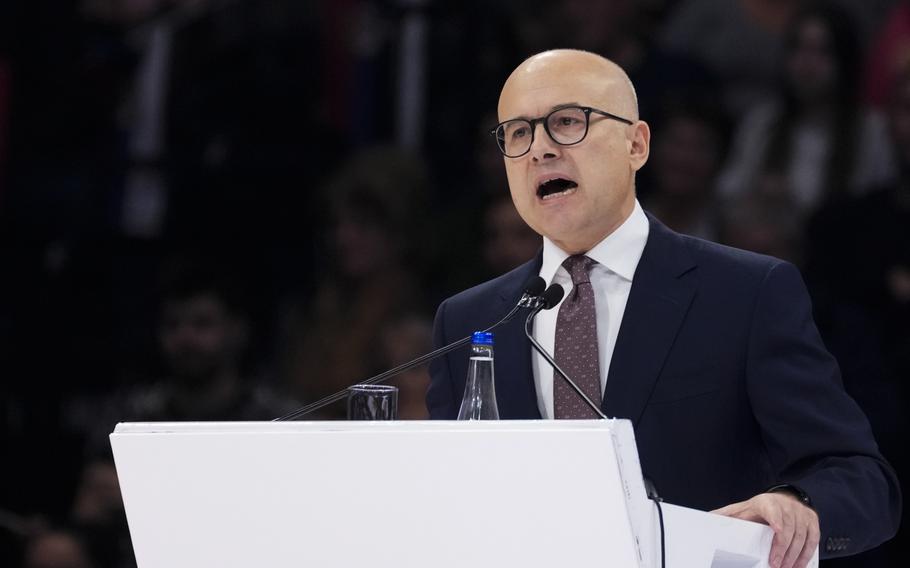 A bald man with a suit and glasses holds a white podium while speaking.