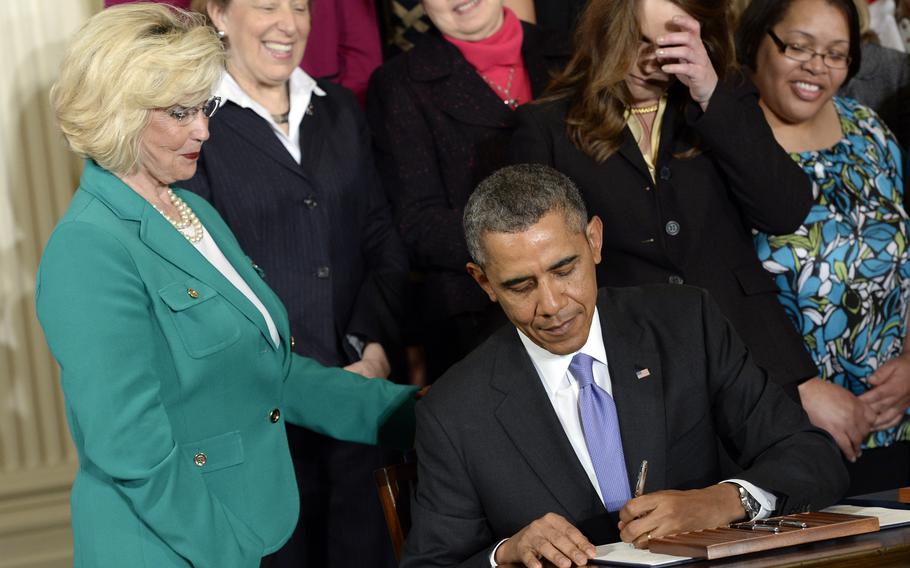 Lilly Ledbetter, among others, stands next to a seated President Barack Obama as he signs a paper at a desk.