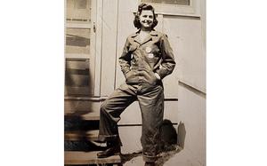 A woman in World War II-era work coveralls poses with one foot resting on a step.